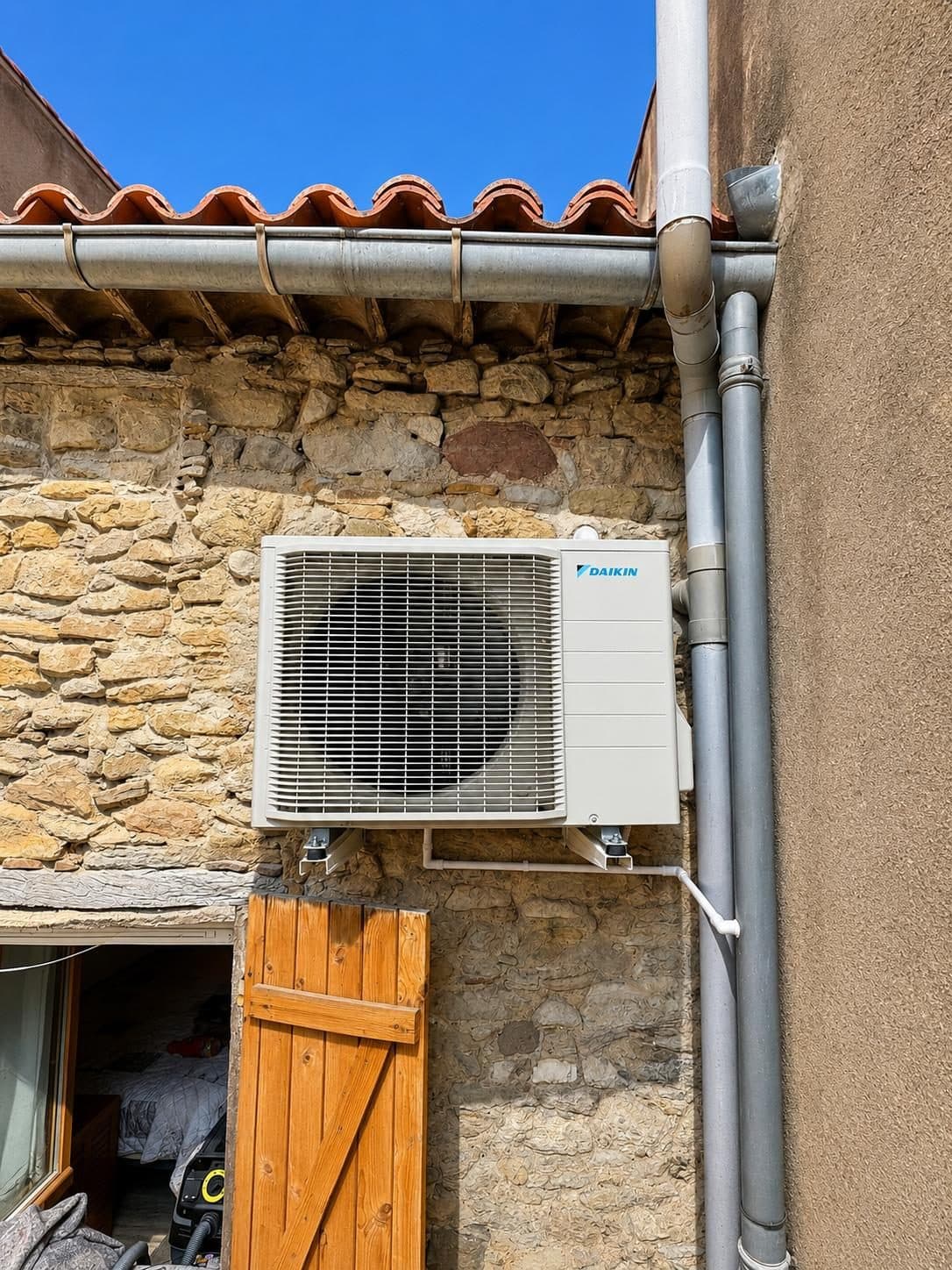 Groupe extérieur de climatisation Daikin fixé sur le mur en pierre d’une maison ancienne à toiture en tuiles canal